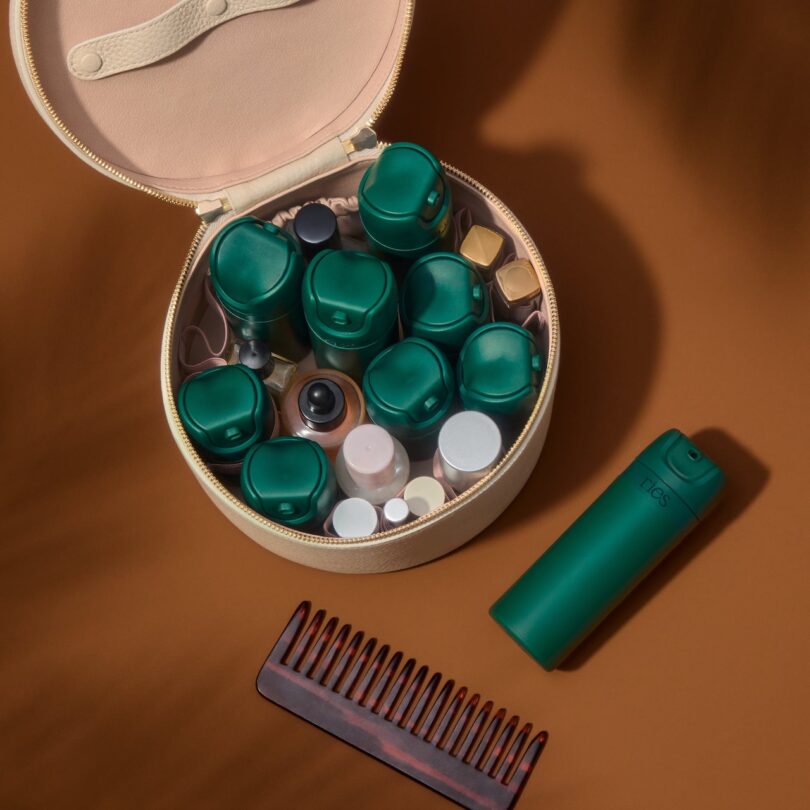 A round travel case filled with various green and white cosmetic bottles is open on a brown surface. Beside it lies a green spray bottle and a brown comb