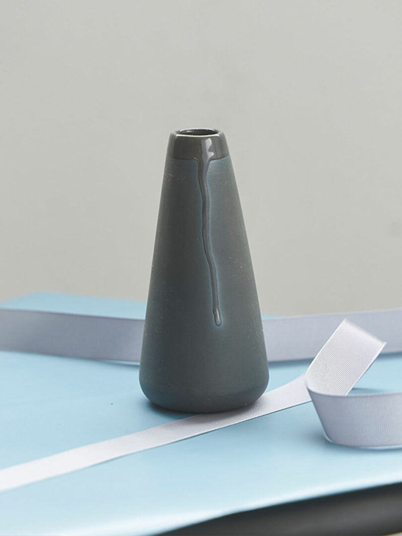 A gray ceramic cone-shaped vase sits on a blue surface with a white ribbon beside it.