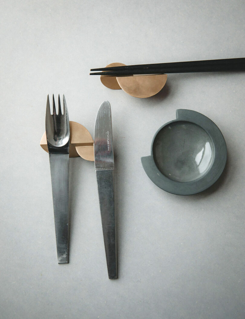 Fork, knife, chopsticks, and a small round dish arranged on a light gray surface.