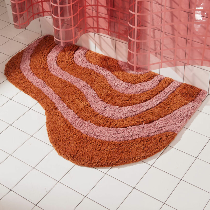 A wavy, pink and orange patterned bath mat on a white tiled floor, next to a semi-transparent red shower curtain.