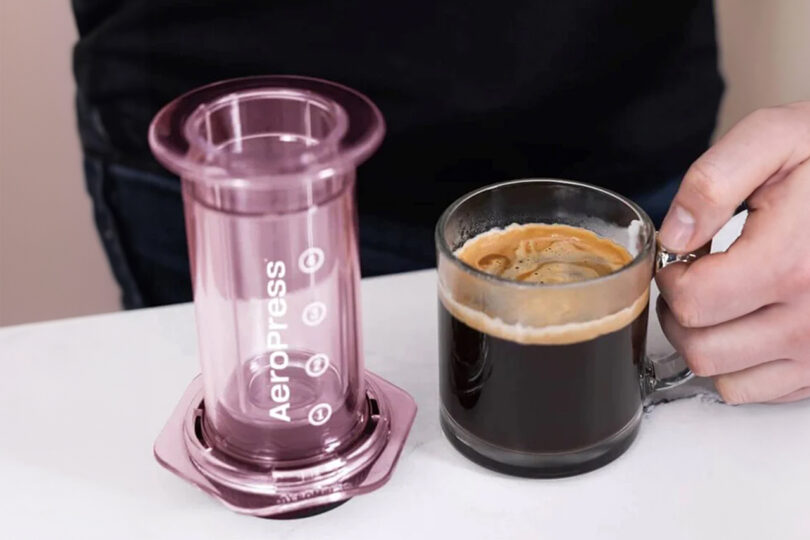 A person holds a glass mug of coffee next to a purple AeroPress coffee maker on a countertop.