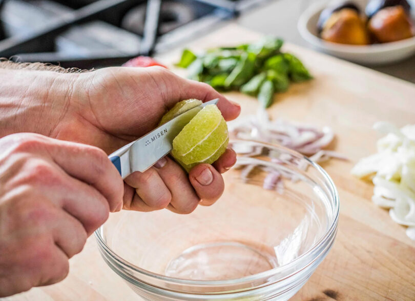 Person using a knife to peel a lime over a glass bowl on a wooden cutting board, with chopped vegetables in the background.