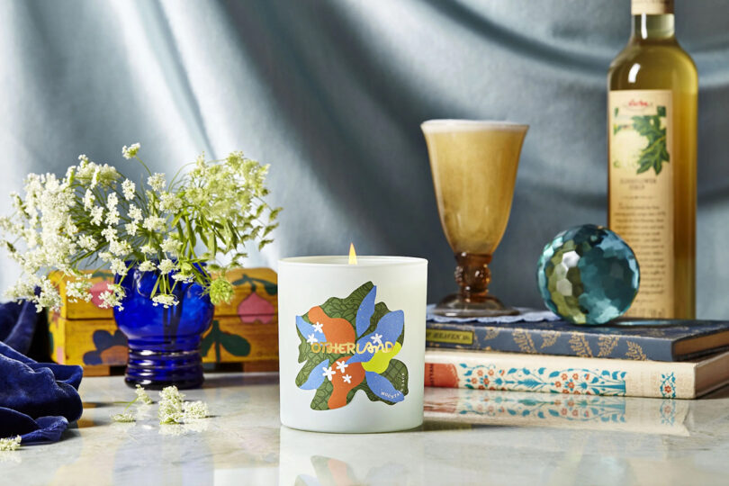 Lit candle on a table with books, a vase of flowers, a drink, a bottle, and decorative items.