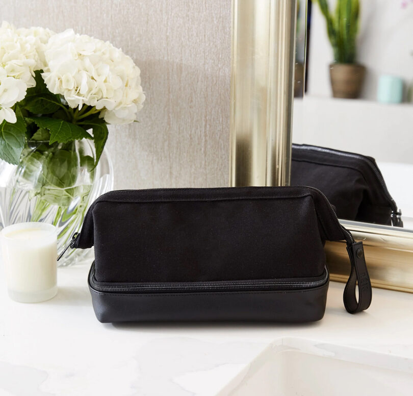 Black toiletry bag on a bathroom counter, next to a vase with white hydrangeas, a candle, and a large mirror.