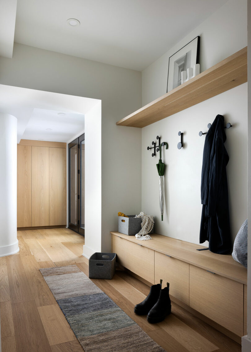 The modern hallway, a nod to Gnome Architects, features wooden flooring, a bench with storage, and coat hooks adorned with a jacket and umbrella. A shelf showcases tasteful decor alongside a cozy rug, all elegantly leading to the next room.