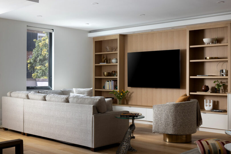 A modern living room features a beige sectional sofa, a patterned armchair, a flat-screen TV on a wooden built-in shelf, and a window offering a serene view of trees.