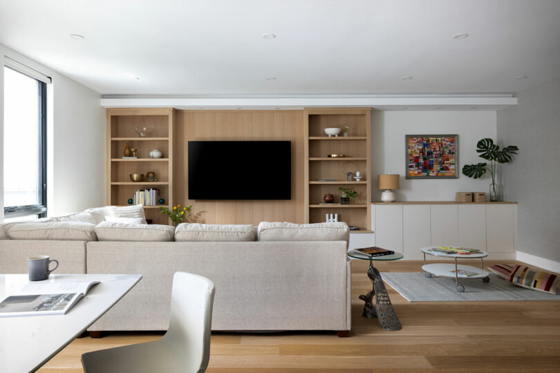 In this modern living room you'll find a beige sectional sofa, wall-mounted TV, and wooden shelves. A coffee table adorned with magazines and a plant adds flair. Light wood flooring and a large window ensure the space feels bright and airy.