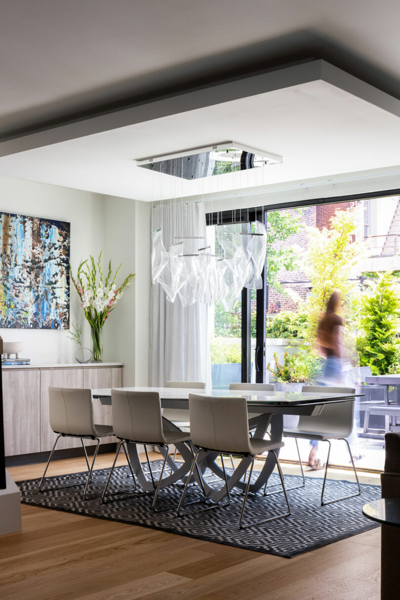 The modern dining room features a white table, six chairs, and a glass chandelier. A large window reveals the enchanting garden beyond. A blurred figure strolls by, while artwork and flowers grace the sideboard.