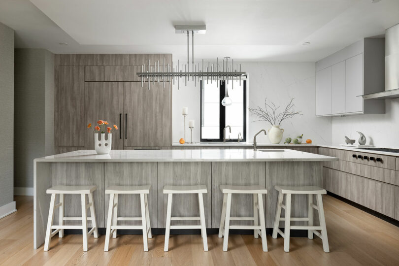 The kitchen features a modern design with a large island, four white stools, and a marble countertop. A chandelier illuminates gray wood cabinets, while a vase of flowers and a bowl of oranges add charm to the inviting space.