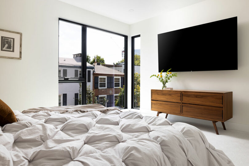 The bedroom features a large bed, a wooden dresser, and a flat-screen TV on the wall. Two large windows offer views of the surrounding buildings.