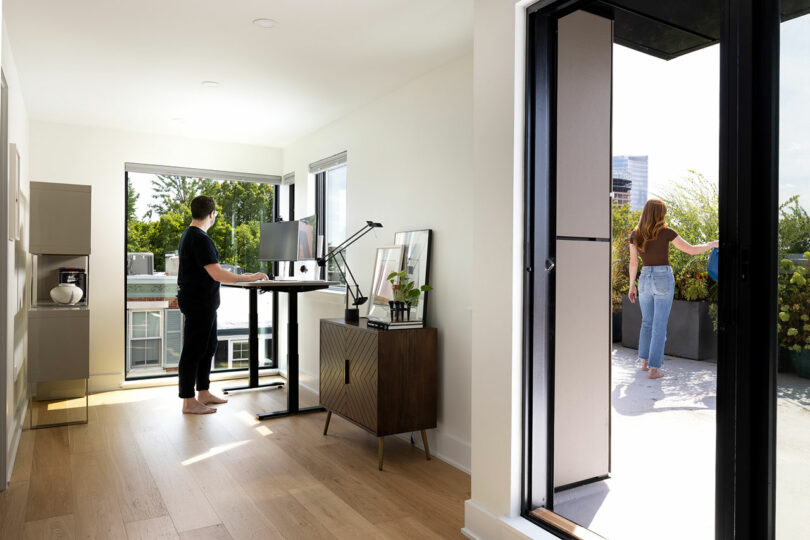 A person stands at a standing desk in a bright room while another enjoys the balcony. The interior boasts light wood floors and modern furnishings that seamlessly blend functionality and whimsy.