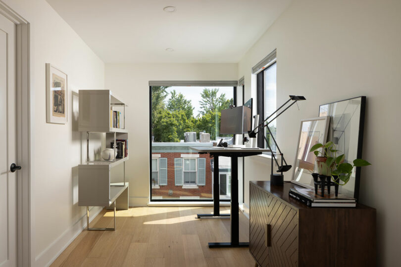 A minimalist home office with a standing desk, monitor, bookshelf, and potted plant. A large window reveals a view of trees and neighboring brick buildings. Neutral color palette prevails.