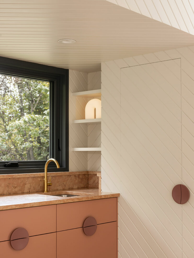 Modern kitchen corner with pink cabinets, circular handles, marble countertop, and a brass faucet. Features a window with a view of trees, and white chevron-patterned walls with shelf and decor.