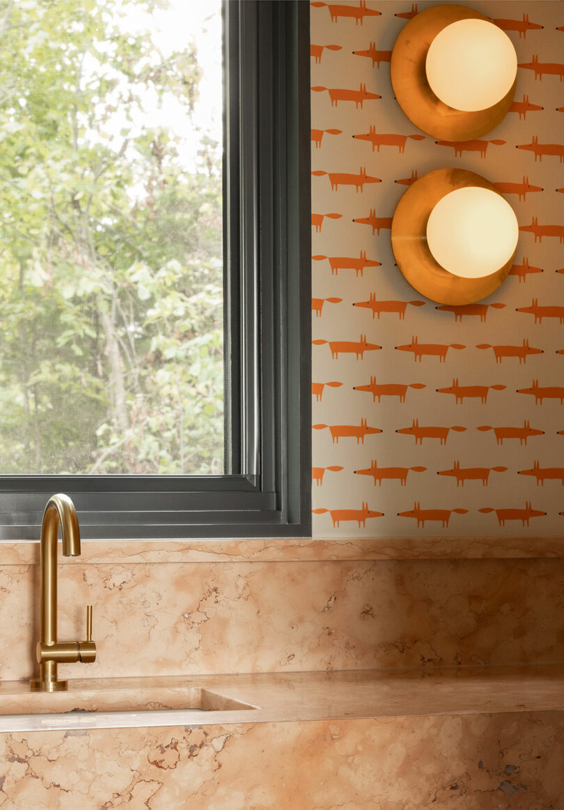 Modern kitchen corner with a marble countertop, gold faucet, window view of greenery, and two spherical wall lights on orange patterned wallpaper.