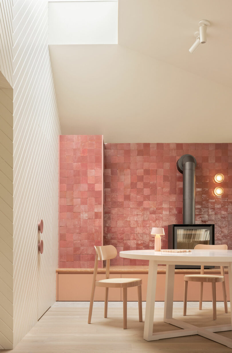 Modern dining area with a white table, wooden chairs, pink tiled wall, wood-burning stove, and soft lighting. The space features a slanted ceiling with a skylight.