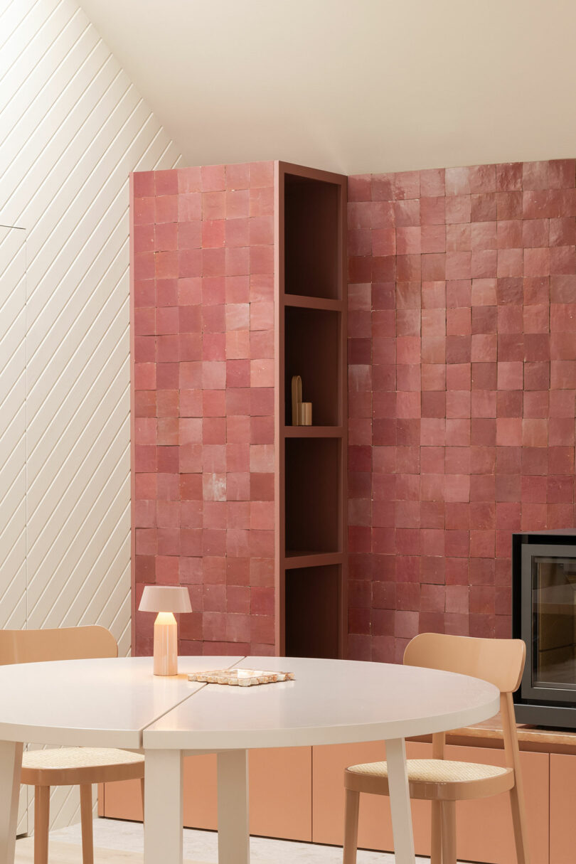 A modern living space featuring a white round table, wooden chairs, a small lamp, and a pink tiled wall with shelves.