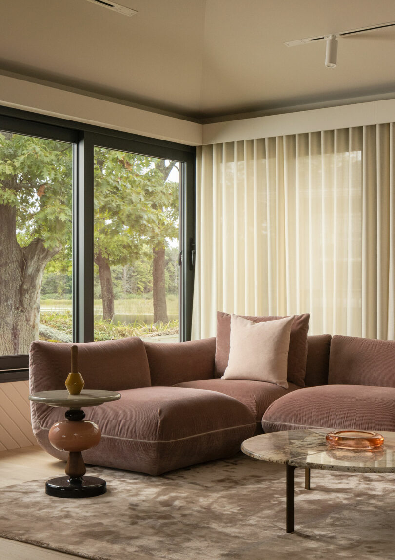 A cozy living room with a pink velvet sectional sofa, marble coffee table, and large windows with a view of trees.