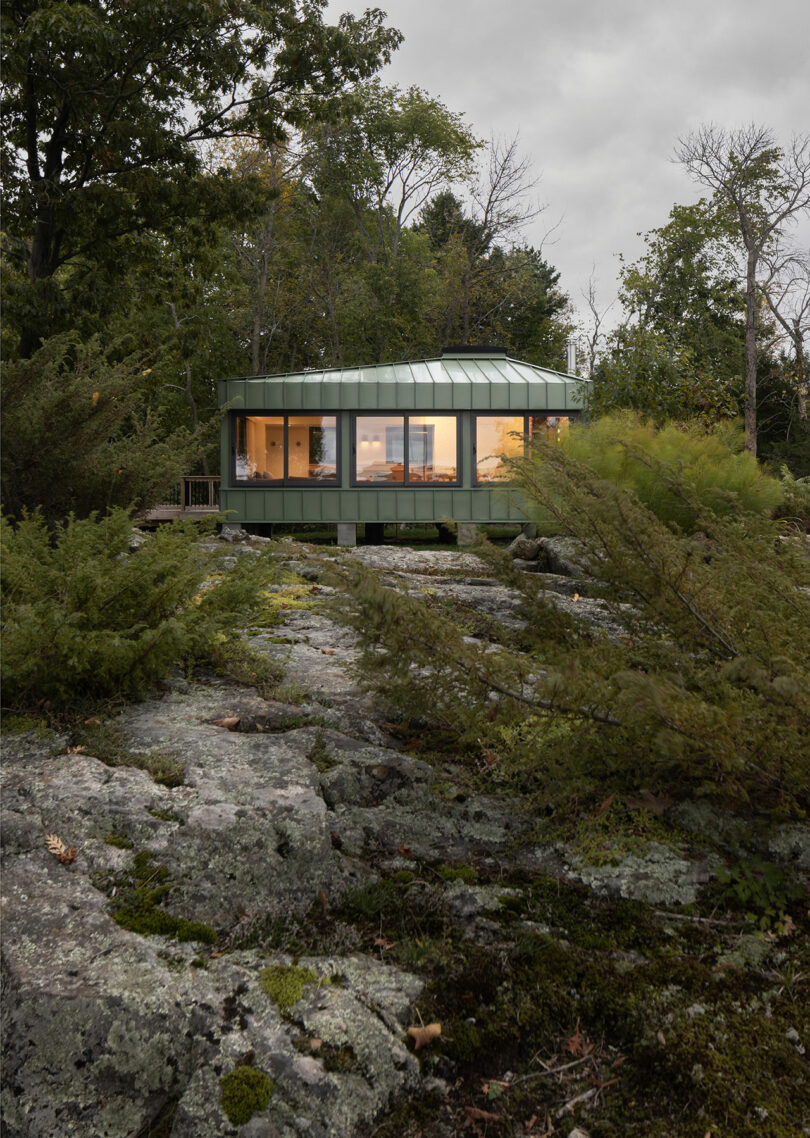 A round, modern green house with large windows is nestled among trees and rocks. It is elevated and surrounded by greenery on an overcast day.