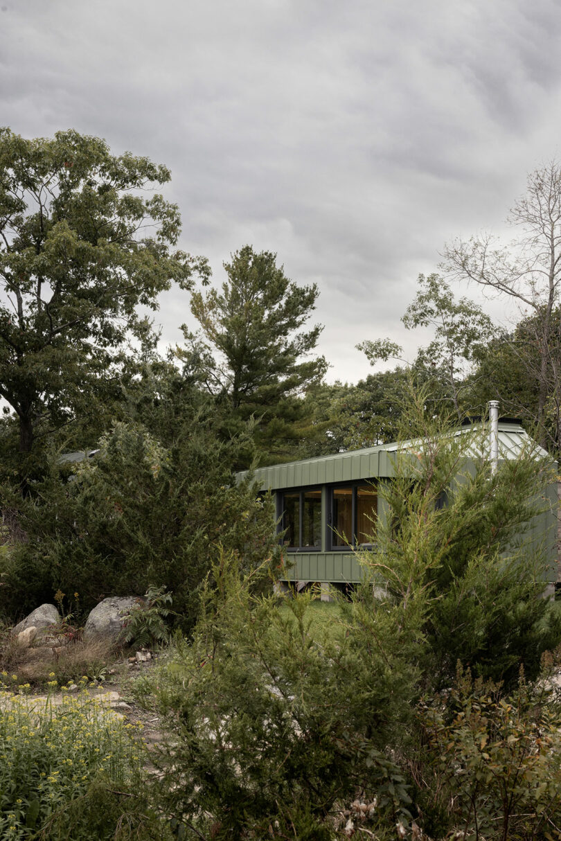 Une petite maison verte entourée d'arbres et de buissons denses sous un ciel nuageux.