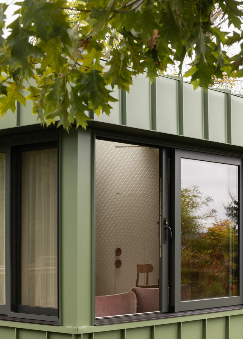 Green exterior wall with large window partially open, revealing a glimpse of a wooden chair and brown sofa inside. Leaves hang in the foreground.