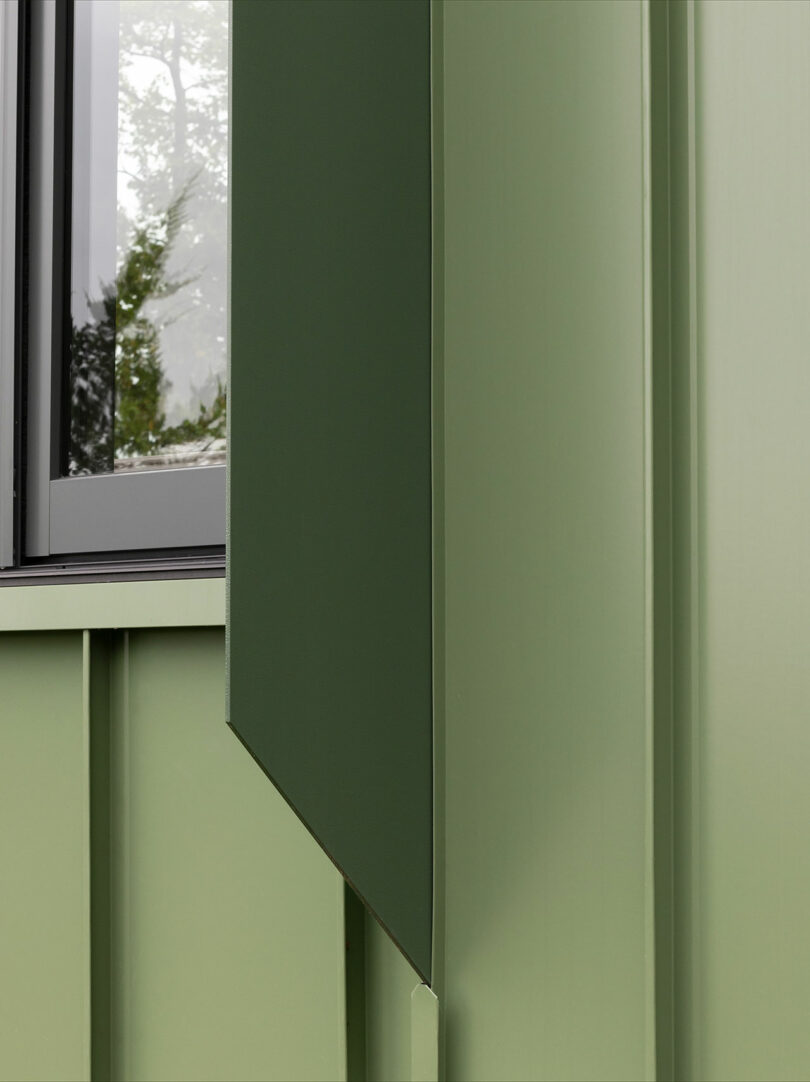 Close-up of a green metal exterior wall with vertical panels, featuring a window with a dark frame. The reflection in the glass shows tree leaves and sky.