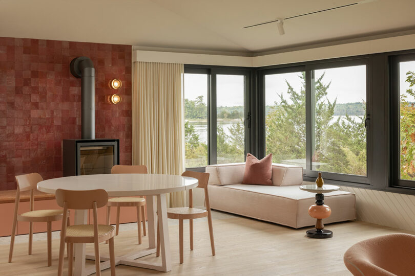 Salon moderne avec une table à manger ronde, des chaises, un poêle à bois et un lit de repos près de grandes fenêtres donnant sur la verdure et l'eau.