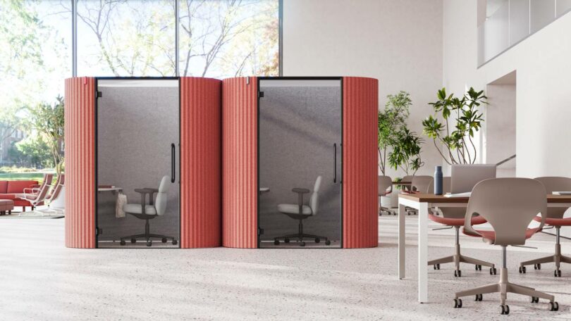 Two red office pods with glass doors and chairs inside, situated in a modern office space with plants and tables.