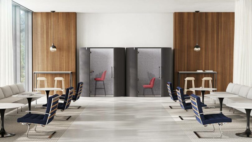 A modern office lounge with blue chairs and small tables, wood-paneled walls, large windows, and two phone booths with red stools in the back.