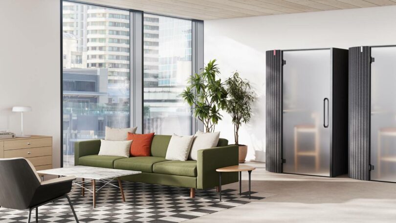 Modern office lounge with a green sofa, assorted cushions, potted plants, a black chair, and two privacy booths near large windows overlooking urban buildings.