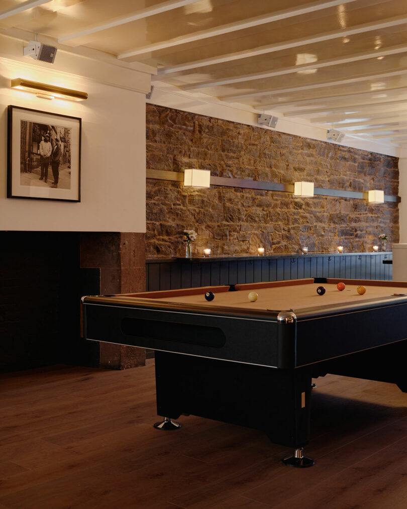 The table in the pool in the room with stone walls and wooden floor, which contains a framed photo on the wall and modern lightning fasteners.