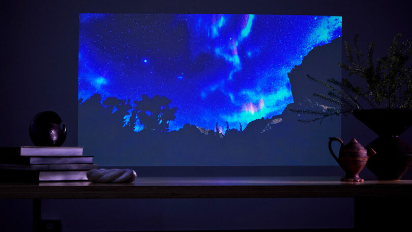 An LG digital projection of a starry night sky and northern lights illuminates the wall, casting a serene glow. In the foreground, a table adorned with books, pottery, and a lush plant completes the tranquil scene.