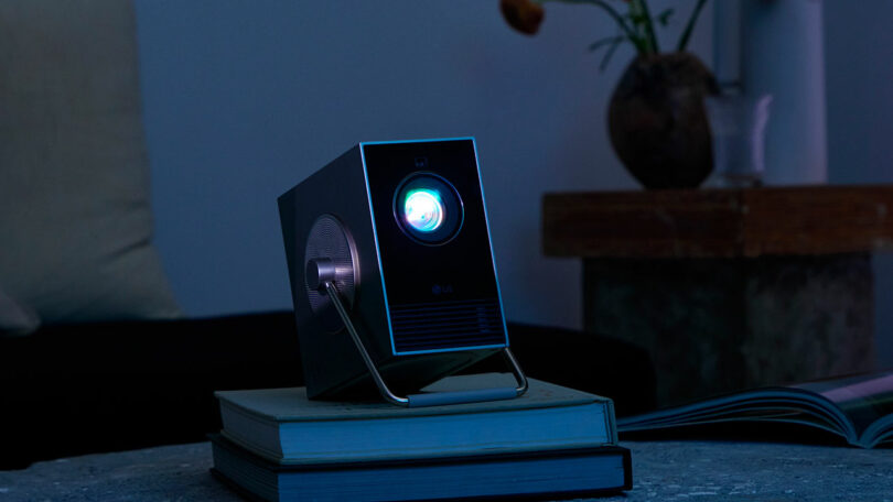 An LG projector emits a blue light while perched on top of two closed books in a dimly lit room.