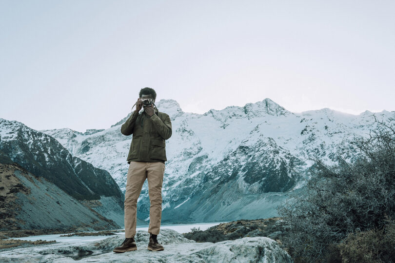 Person in winter clothing stands on a rocky surface, looking through binoculars, with snow-covered mountains and a clear sky in the background.