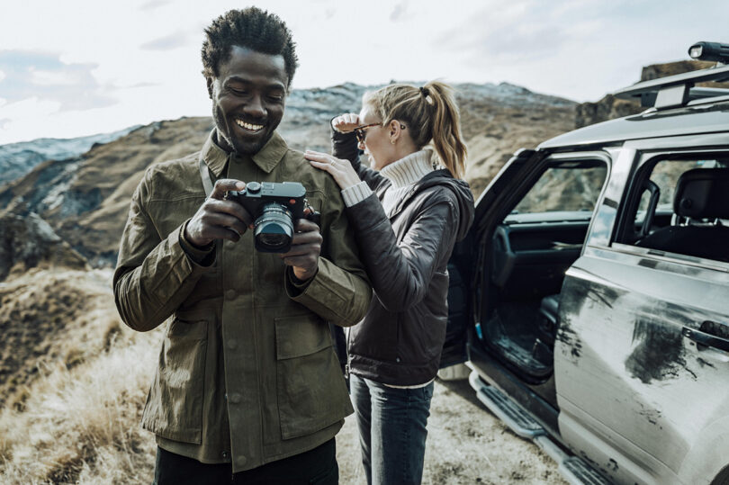 Two people outdoors by a vehicle; one is holding a camera with a smile, while the other is looking into binoculars, mountain landscape in the background.