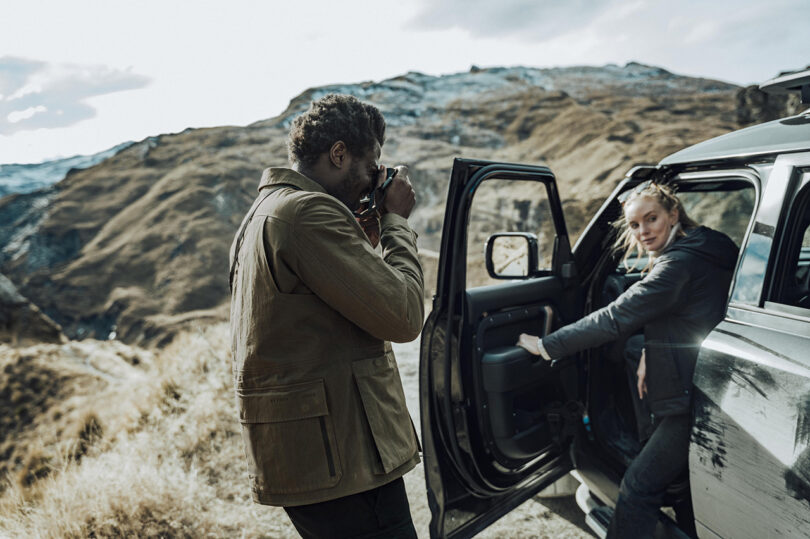 A person takes a photo of another person who is stepping out of a car in a mountainous landscape.