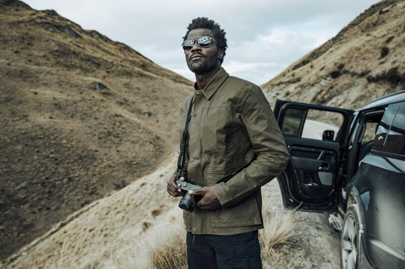 Man standing near an open car door in a mountainous area, holding a camera and wearing sunglasses.