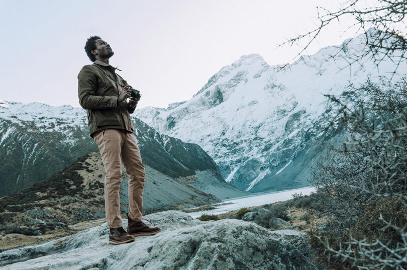 A person stands on a rocky ledge holding a camera, gazing upward in a mountainous landscape with snow-covered peaks in the background.