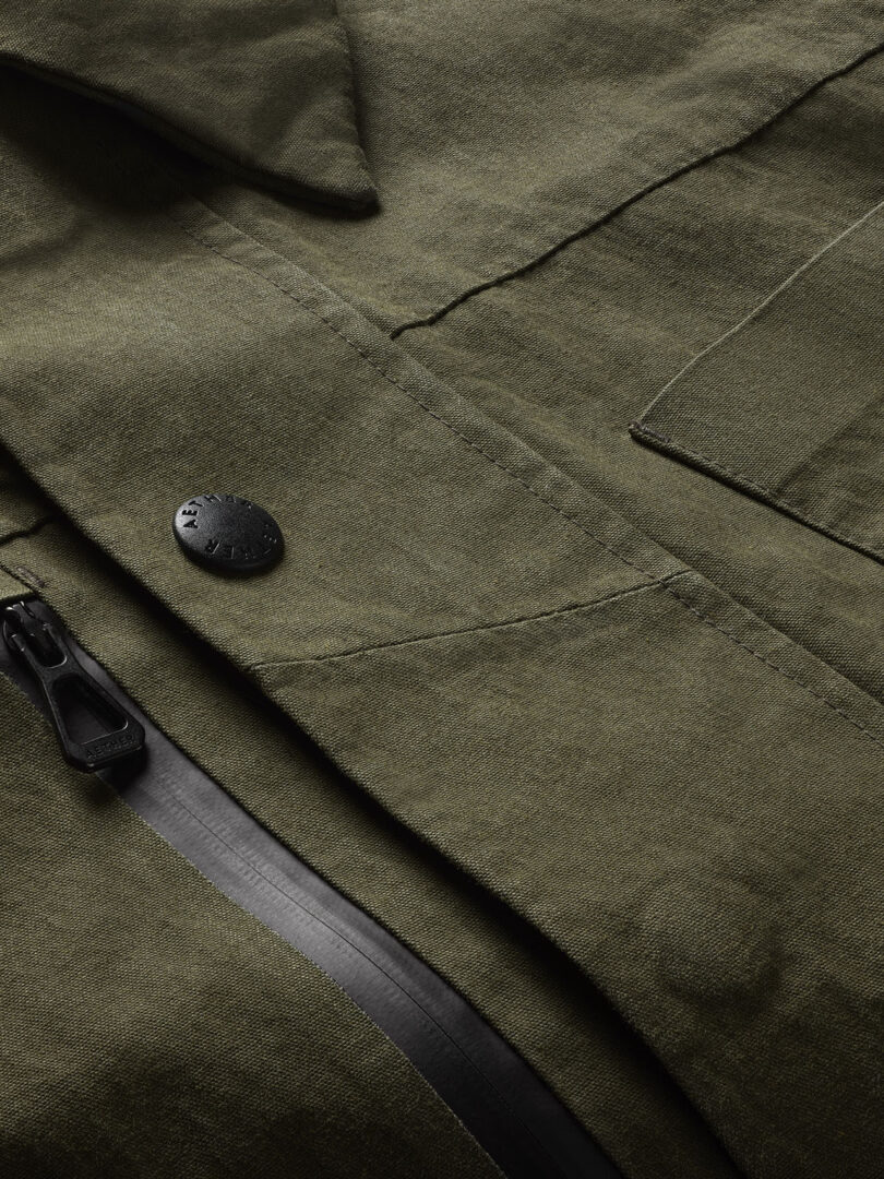 Close-up of an olive green jacket showing a metal button, a zipper, and a pocket flap. The fabric has a textured appearance.