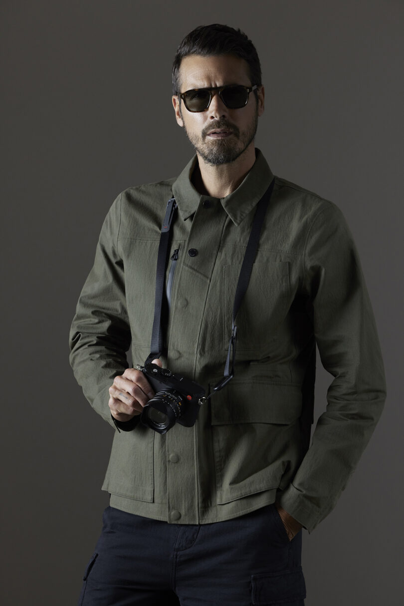 A man wearing sunglasses and a green jacket holds a camera against a neutral background.