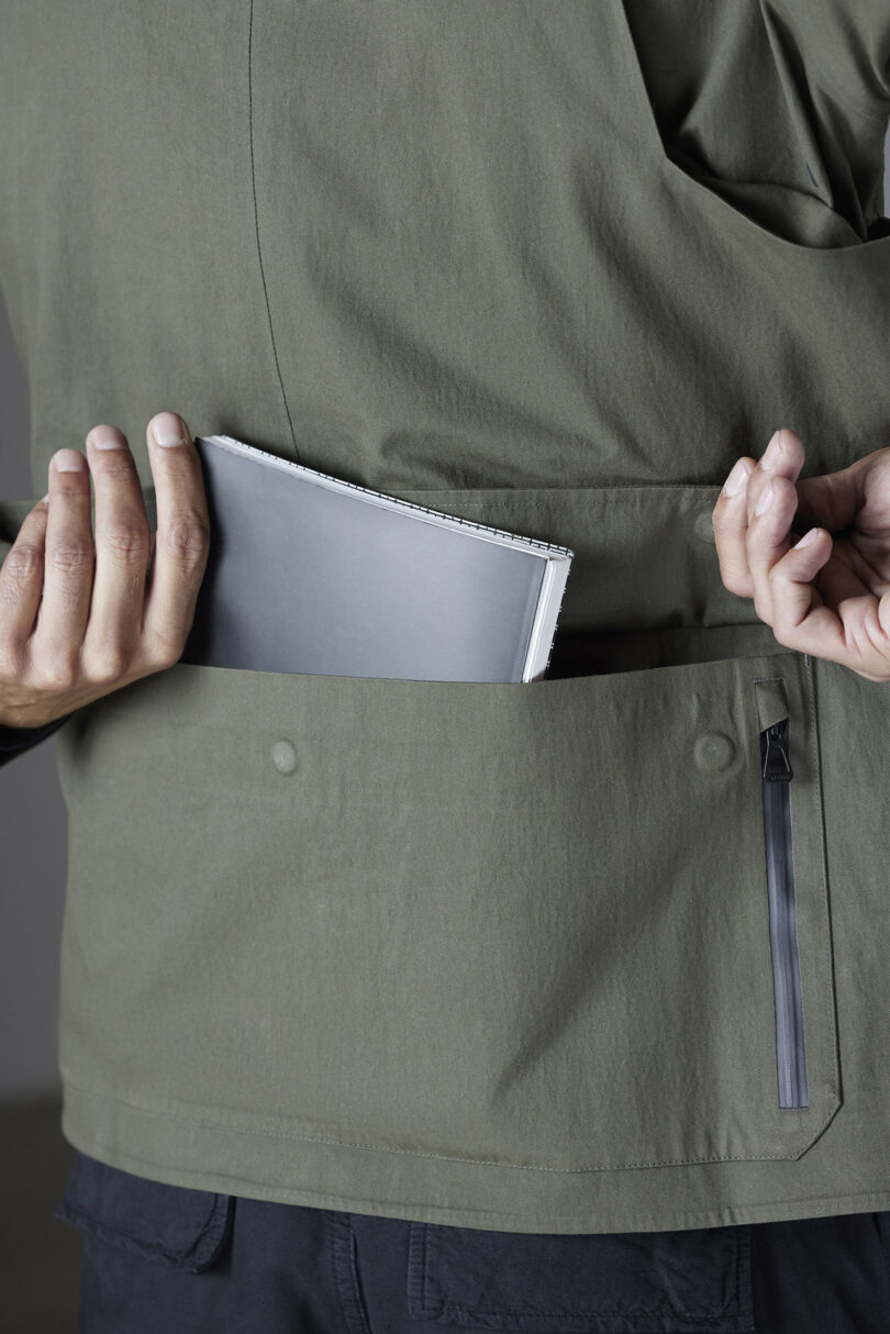 Person placing a notebook into the back pocket of a green jacket.