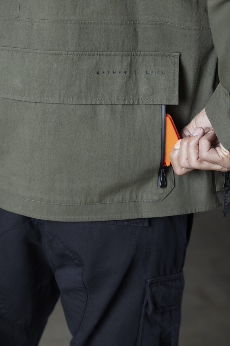 Person placing an orange object into the back pocket of a green jacket wearing black pants.