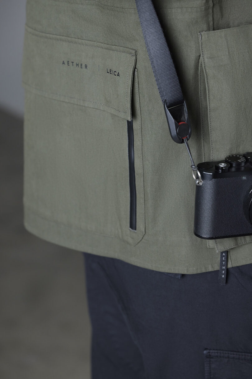 Close-up of a person wearing an olive green AETHER jacket with large pockets, holding a black Leica camera on a strap. The focus is on the jacket's texture and branding.