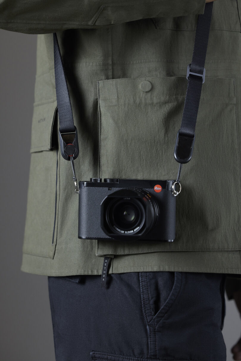 A person wearing a green jacket and dark pants has a black camera with a red logo hanging from a neck strap.