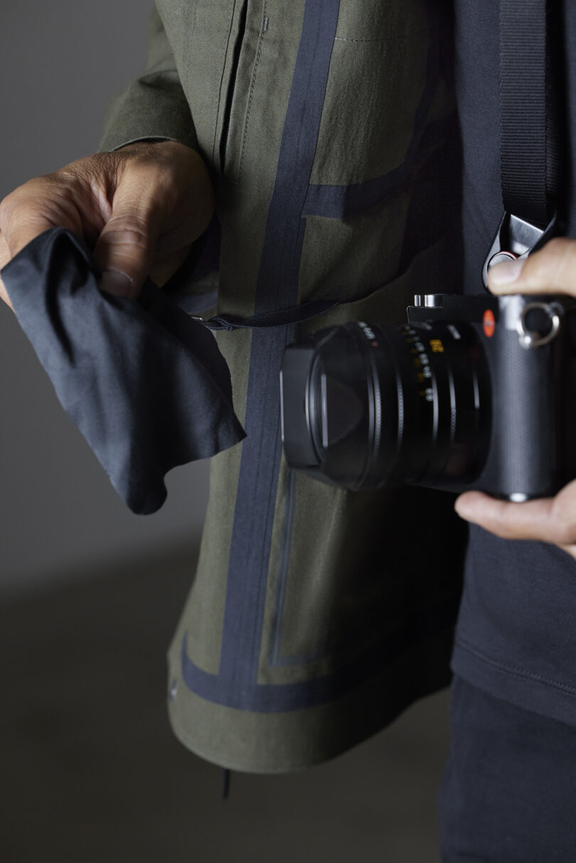 Person holding a black camera in one hand and a lens cloth in the other, wearing a green jacket.