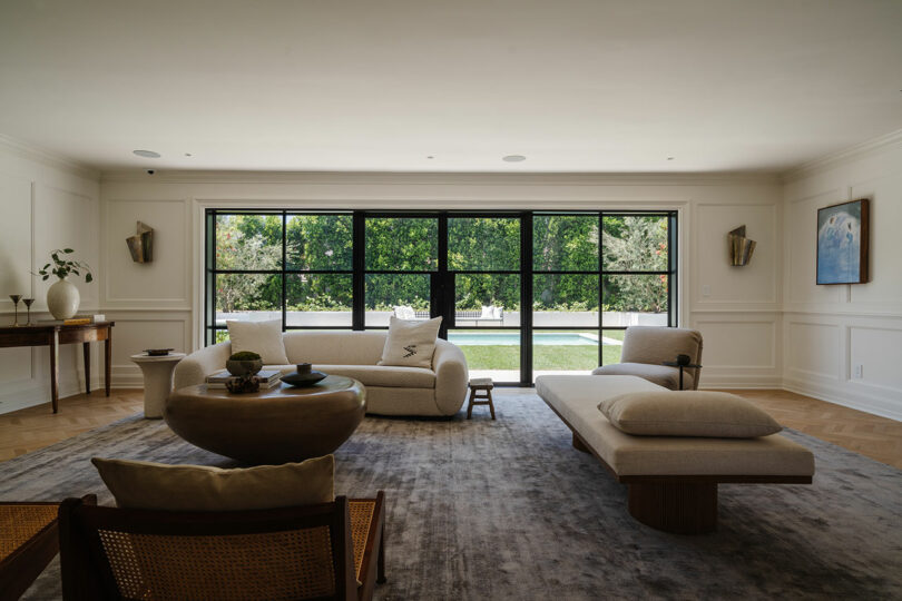 Spacious living room with a large window, featuring a beige sofa, lounge chair, and a wooden table.