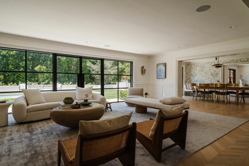 A modern living room featuring large windows, cream sofas, and wooden chairs surrounds a circular coffee table. In the background, the dining area boasts floral wallpaper.