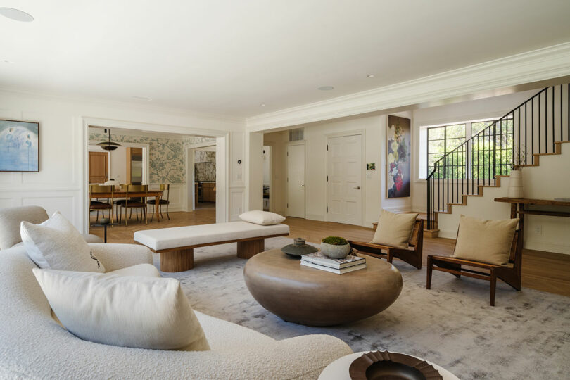 A spacious living room with a curved sofa, wooden coffee table, two chairs, and a view of the dining area and staircase. The light walls and decor create an inviting atmosphere reminiscent of warm hospitality.