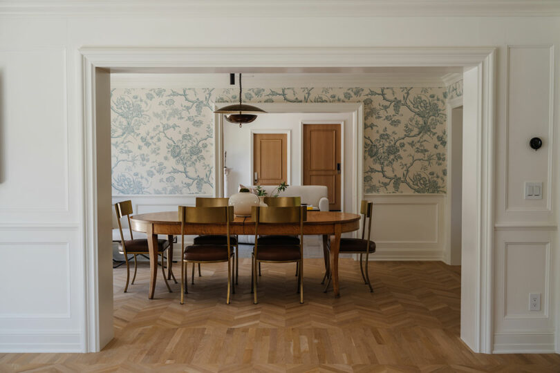 The dining room features a wooden table and chairs, complemented by light wood herringbone floors. Floral wallpaper and white walls create an elegant backdrop, while a pendant light casts a warm glow, completing the inviting ambiance.