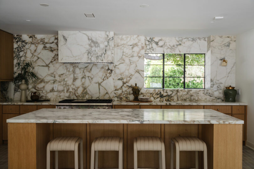 A modern kitchen featuring a marble island and backsplash, wooden cabinets, and four stools awaits. A large window provides views of the lush greenery outside.