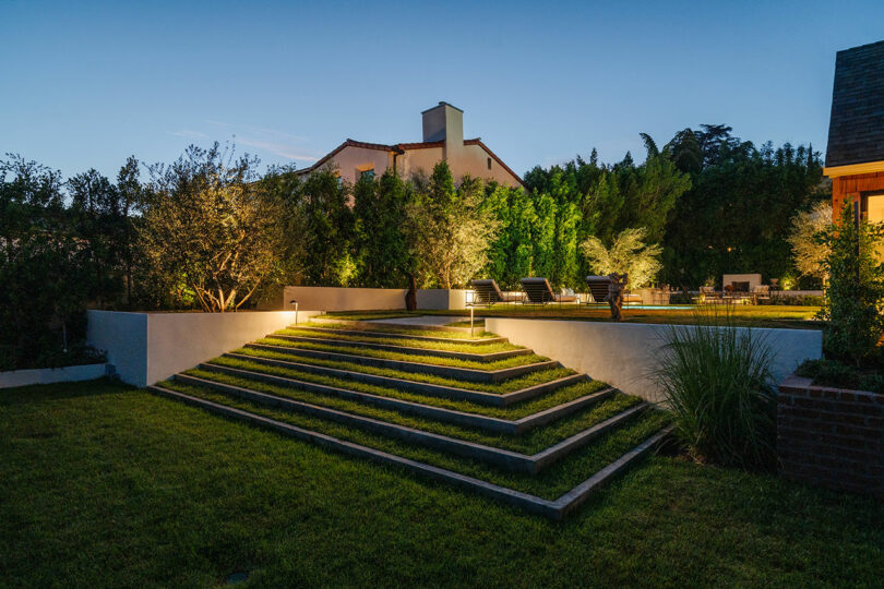 Wide stone steps lead up to a garden area, where trees and lighting create a tranquil ambiance in their backyard at dusk.